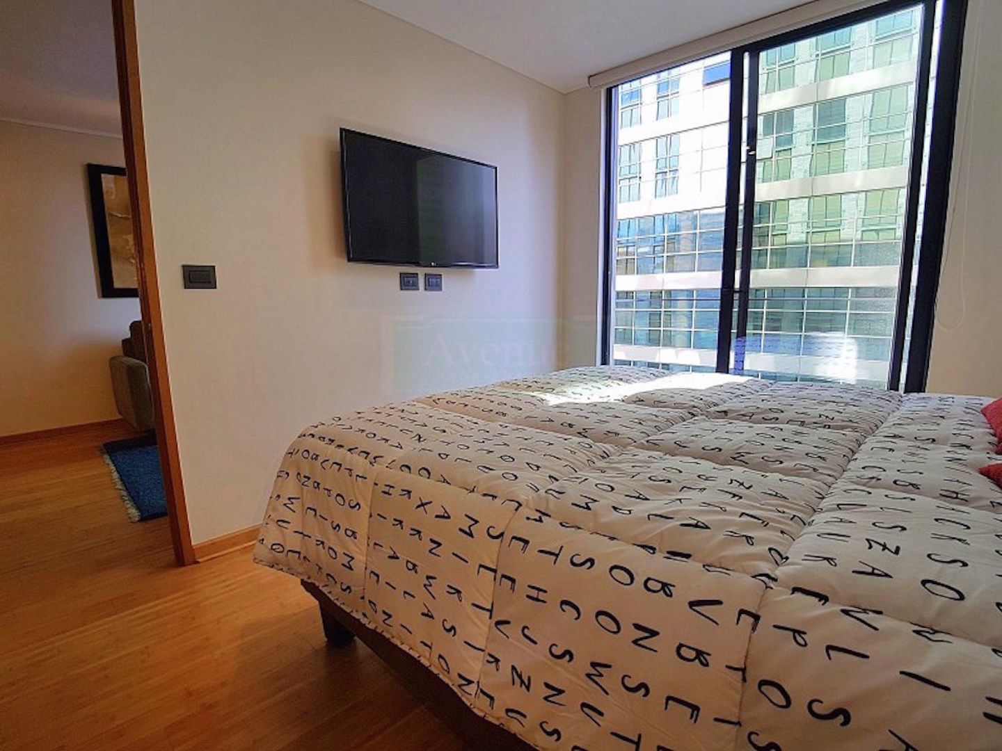 A bedroom with a large window, a flat-screen TV mounted on the wall, and a bed with a patterned bedspread.
