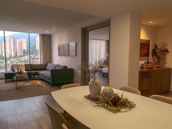 Una sala de estar moderna con sofá, mesa de centro y mesa de comedor. Se ve una vista de edificios altos por la ventana.