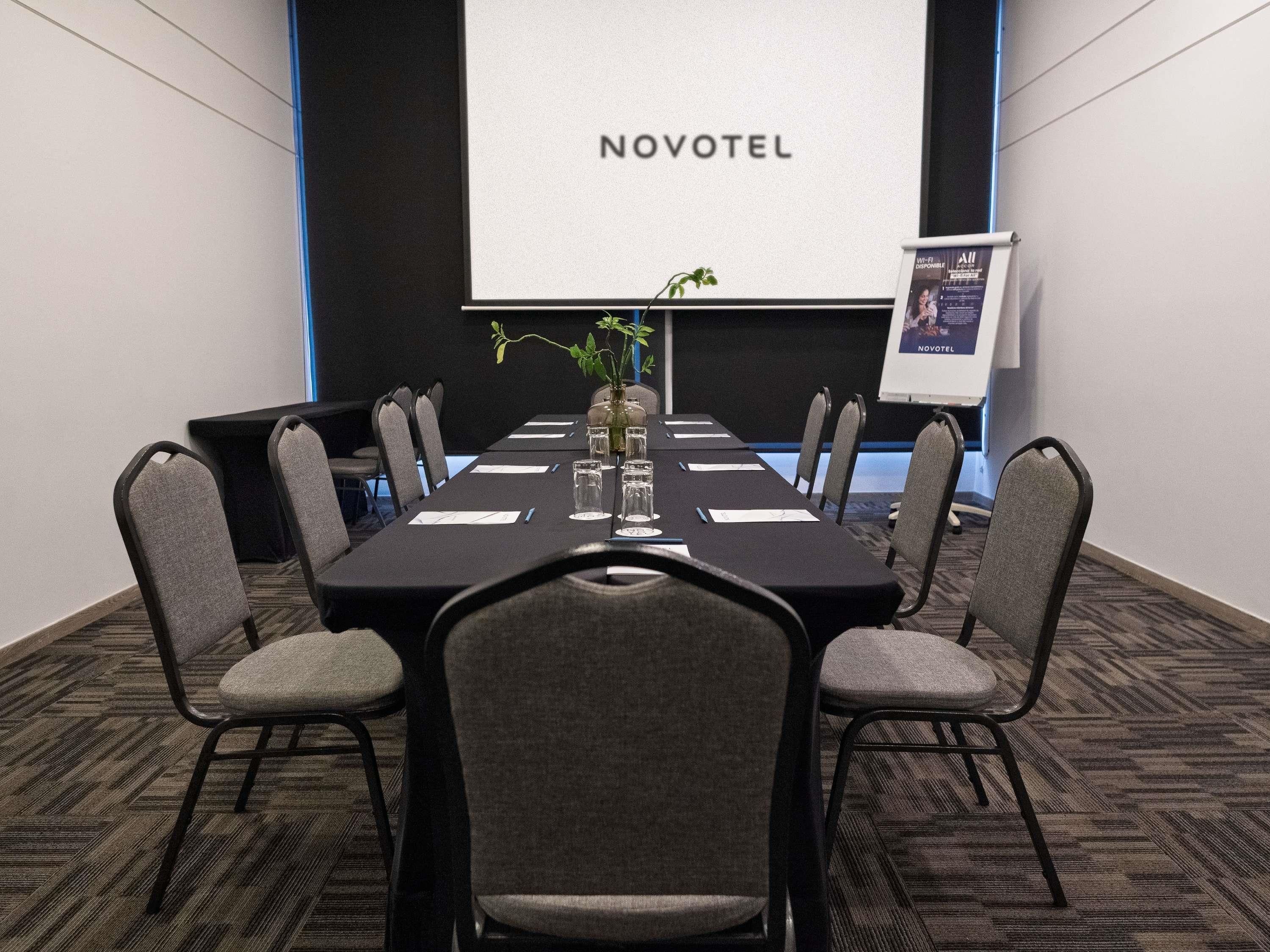 A conference room with a long table, chairs, a projector screen, and a sign reading 'NOVOTEL'.