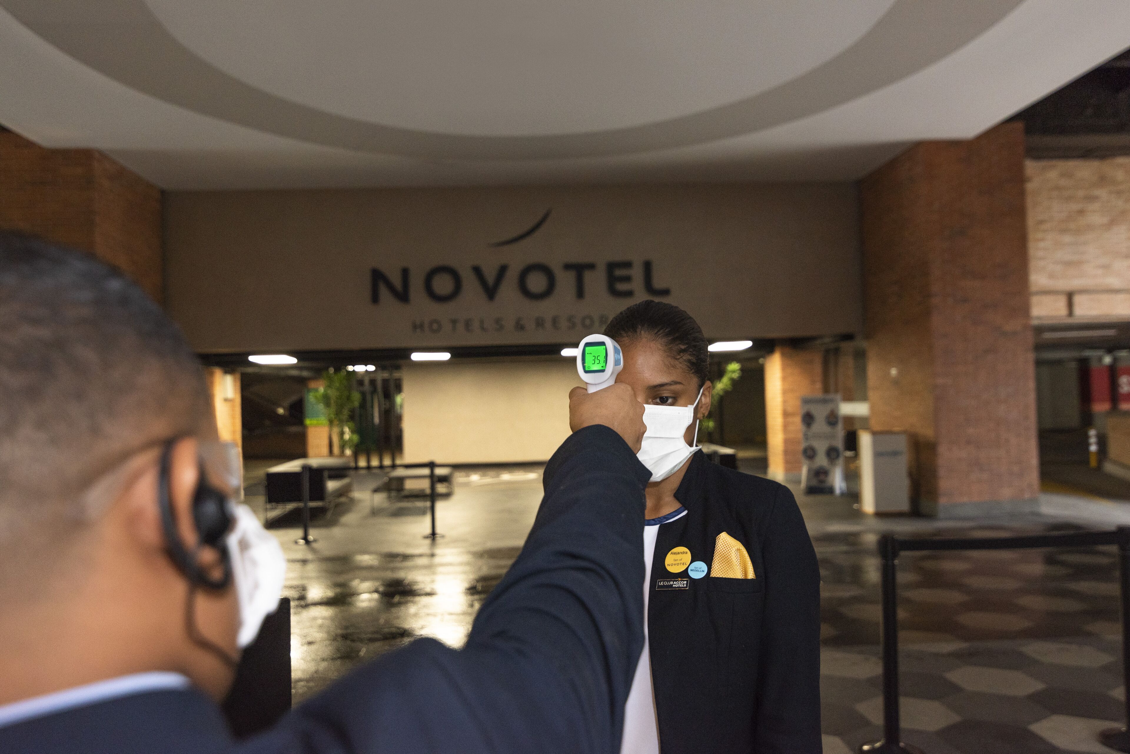 A person wearing a mask and a badge, standing in front of a Novotel hotel entrance.