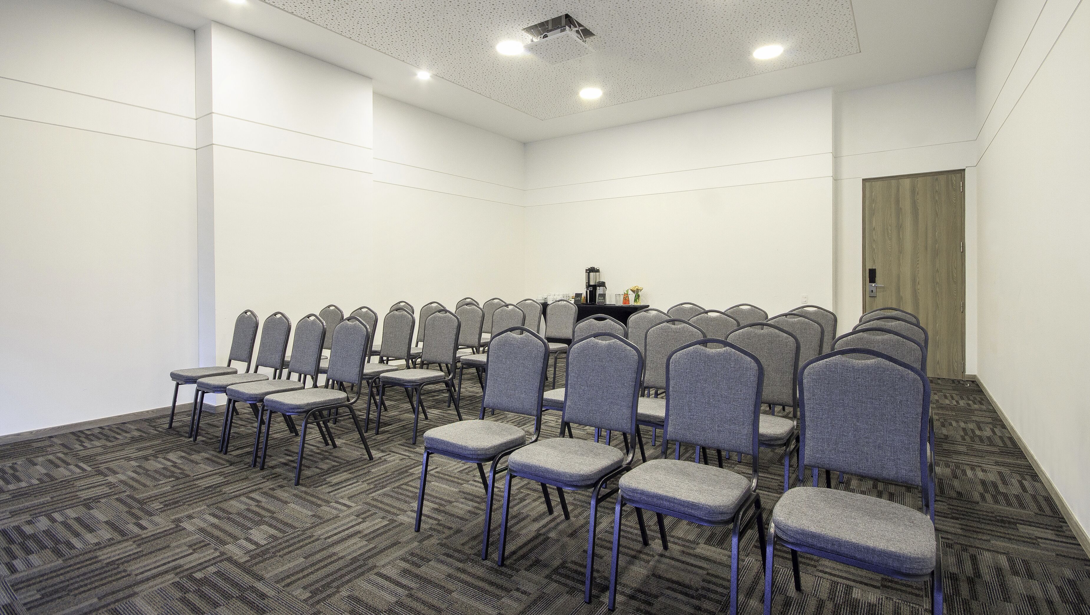 A conference room with rows of grey chairs, a black table with a coffee maker, and a wooden door.