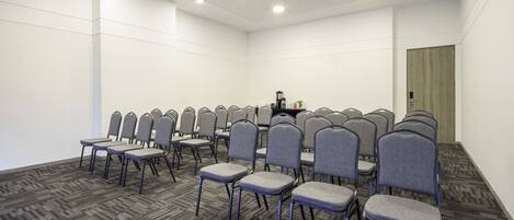 A conference room with rows of grey chairs, a black table with a coffee maker, and a wooden door.