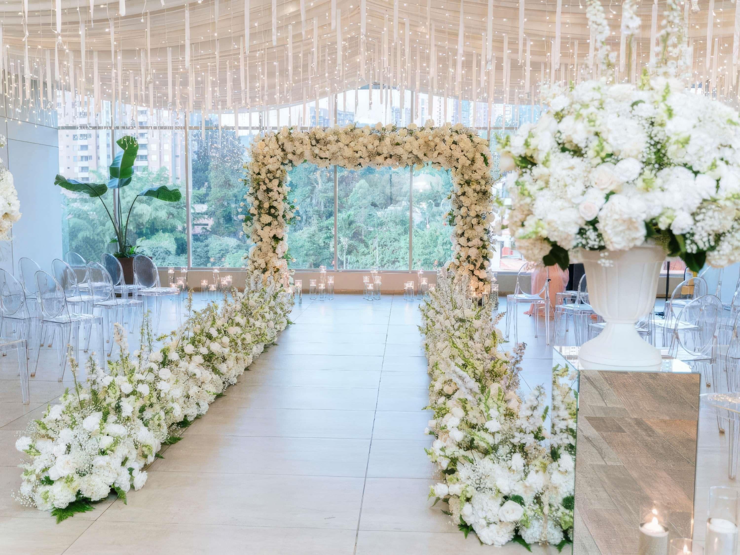 Un espacio interior con un arco floral, sillas transparentes y vistas al verdor exterior.