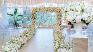 Un espacio interior con un arco floral, sillas transparentes y vistas al verdor exterior.