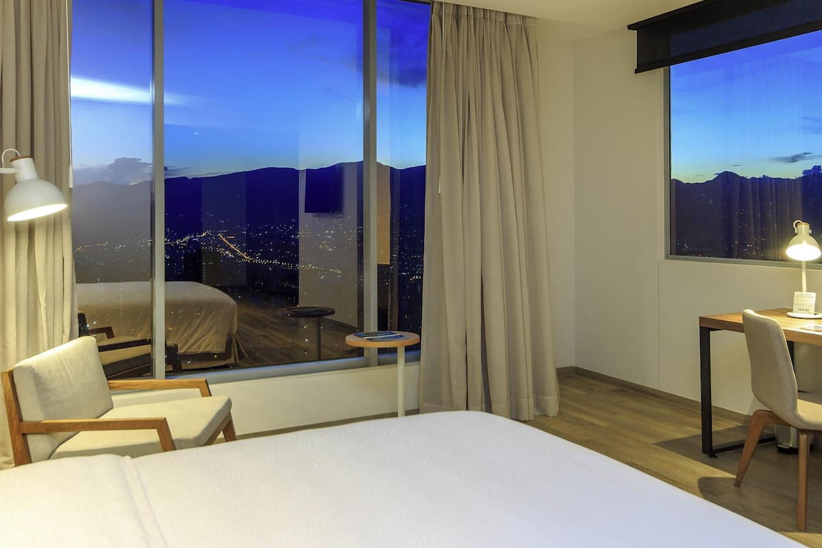 Une chambre d’hôtel avec un grand lit, un bureau avec une chaise et une vue sur le paysage urbain illuminé la nuit.