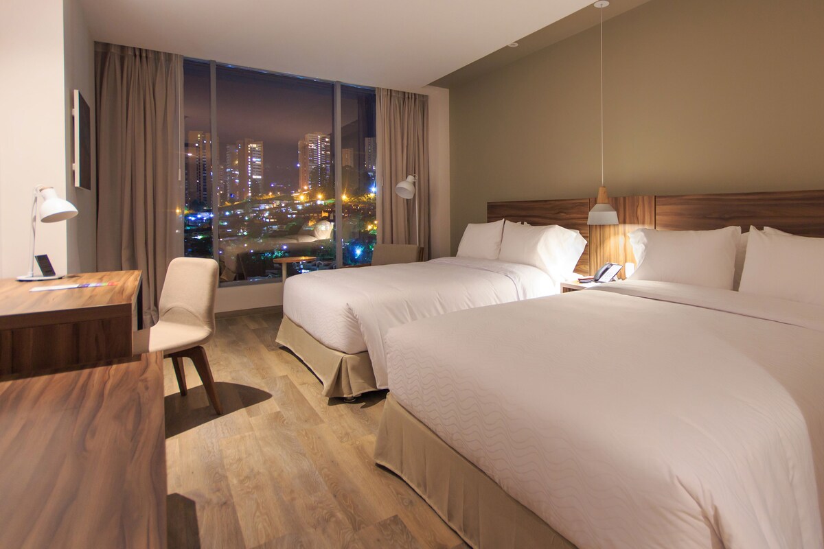 Een hotelkamer met twee bedden, een bureau, een stoel en uitzicht op een stadsbeeld bij nacht.