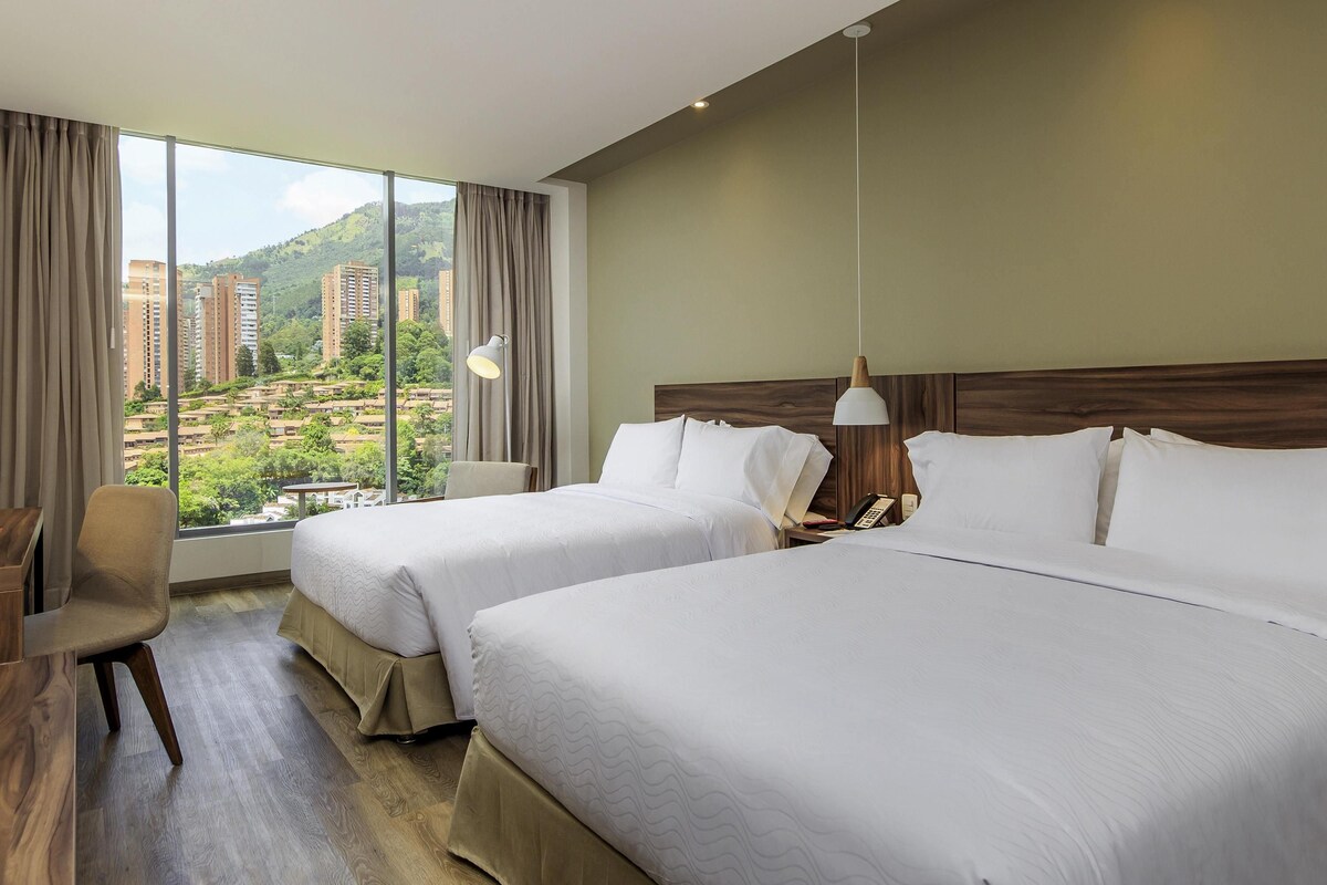 A hotel room with two beds, a desk, and a view of buildings and greenery.
