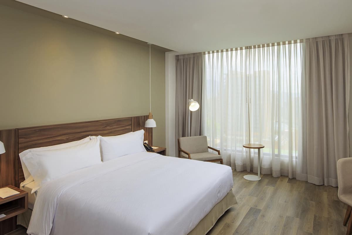 Une chambre d’hôtel avec un grand lit, une chaise, une petite table et une fenêtre avec des rideaux.
