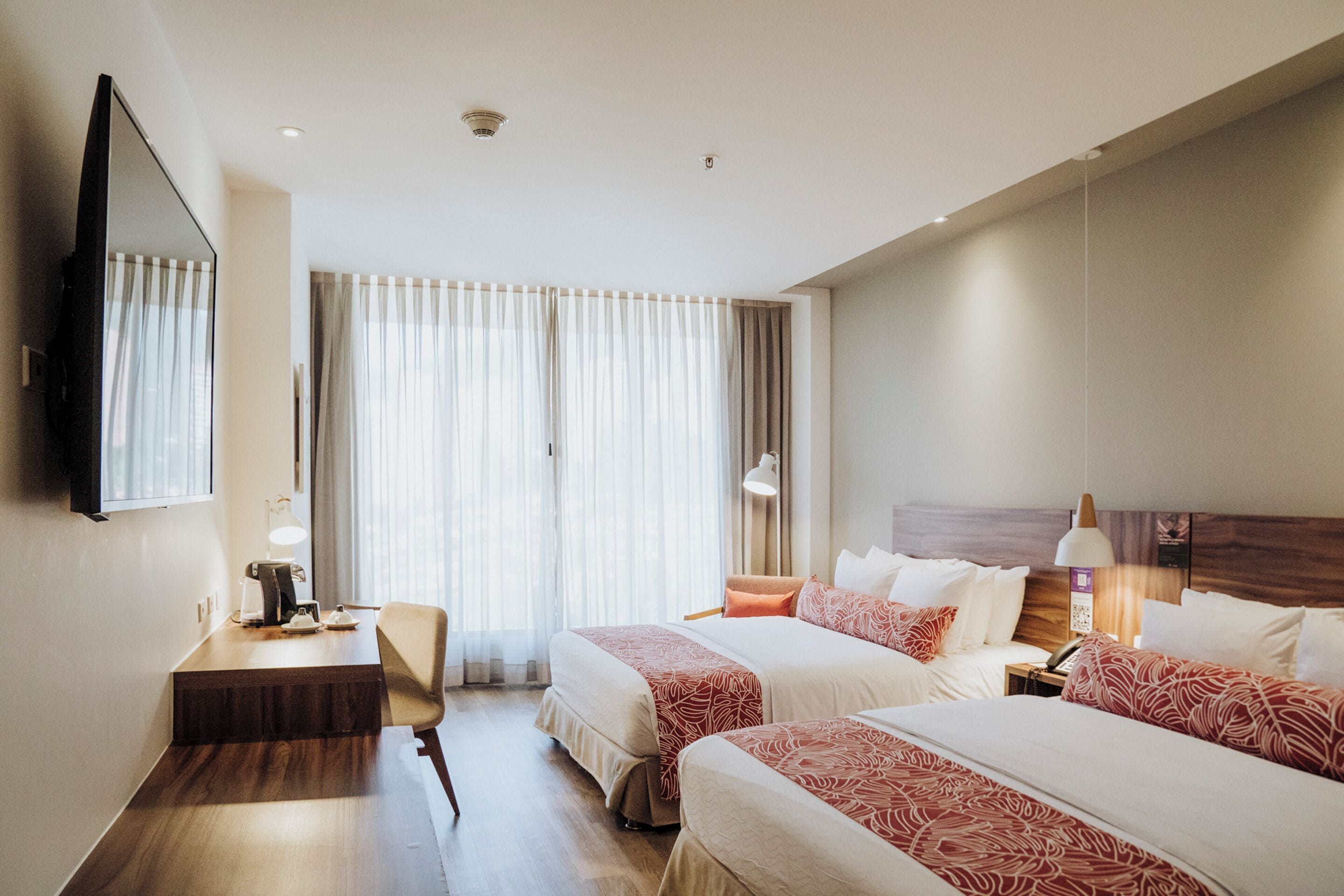 A hotel room with two beds, a desk, a chair, a TV, and a window with curtains.