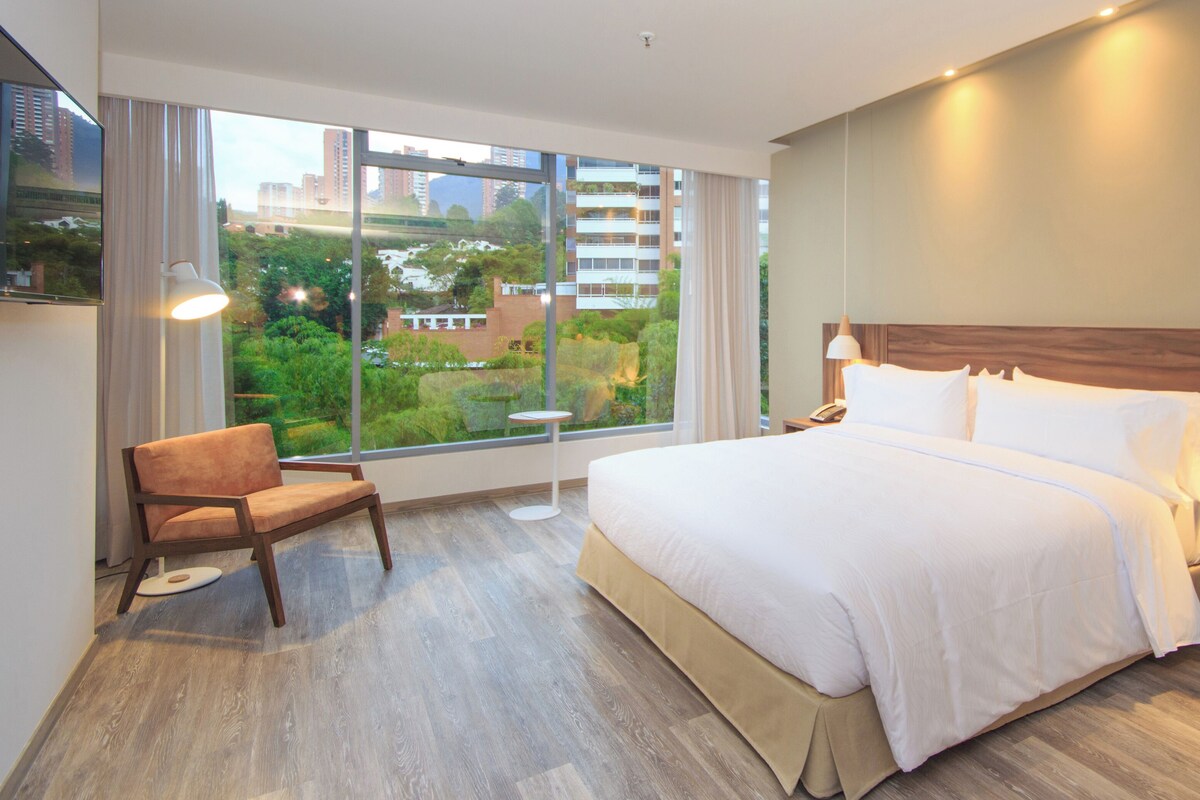 Een moderne hotelkamer met een groot bed, een stoel, een nachtkastje en uitzicht op een stadsgezicht door grote ramen.