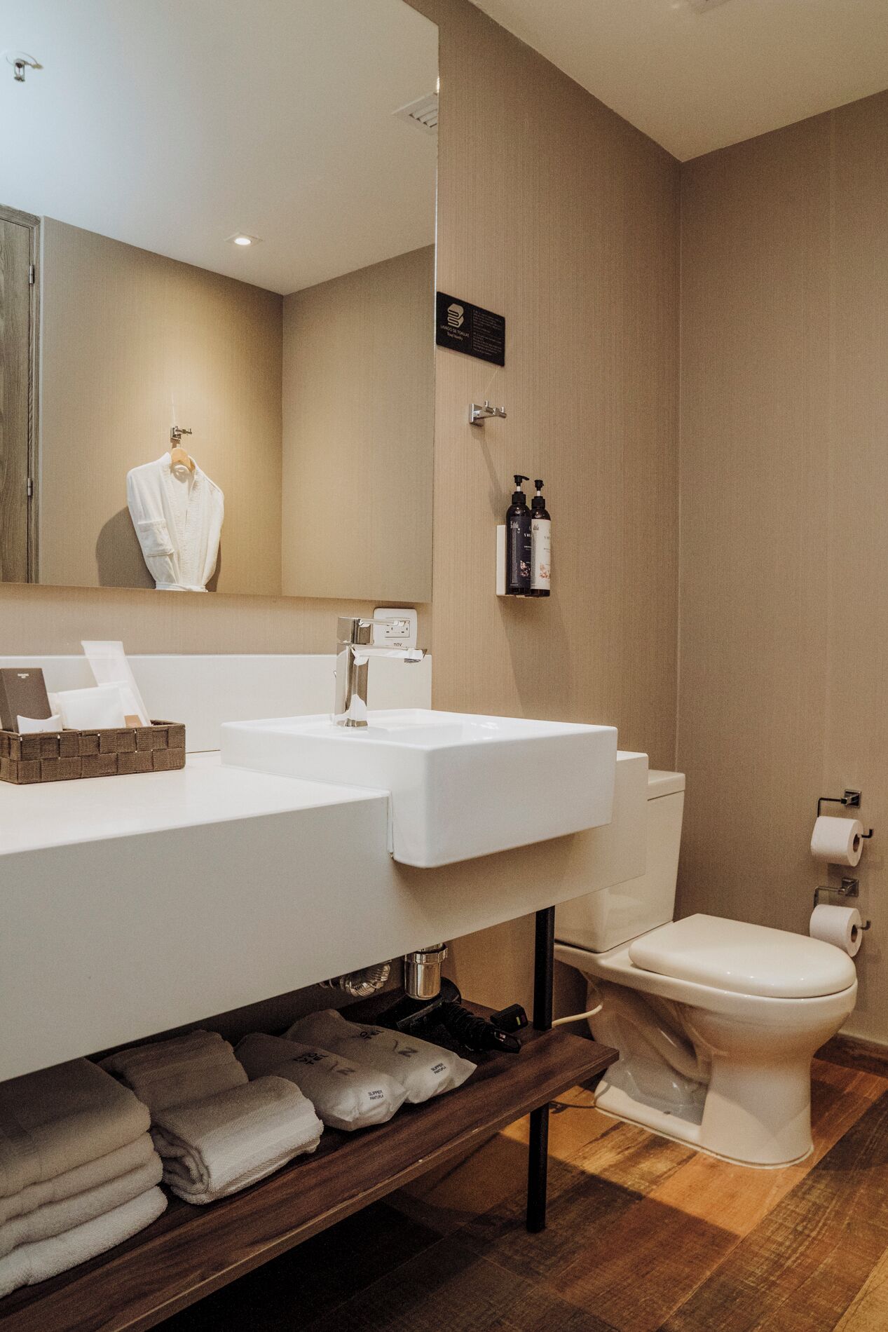 A modern bathroom with a large mirror, two sinks, a toilet, and a shelf with towels.