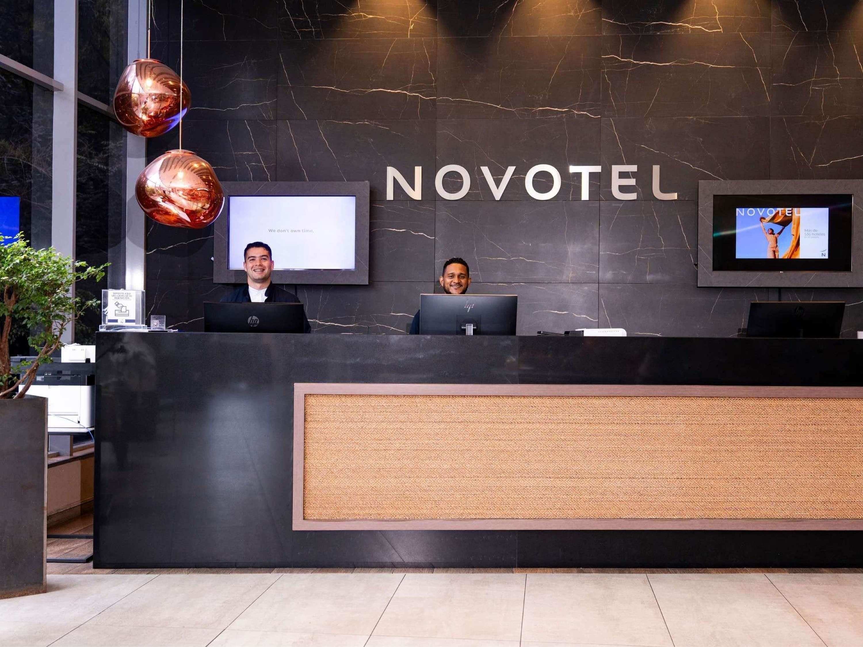 A reception desk with two employees, a marble wall, and a TV screen displaying 'NOVOTEL.'