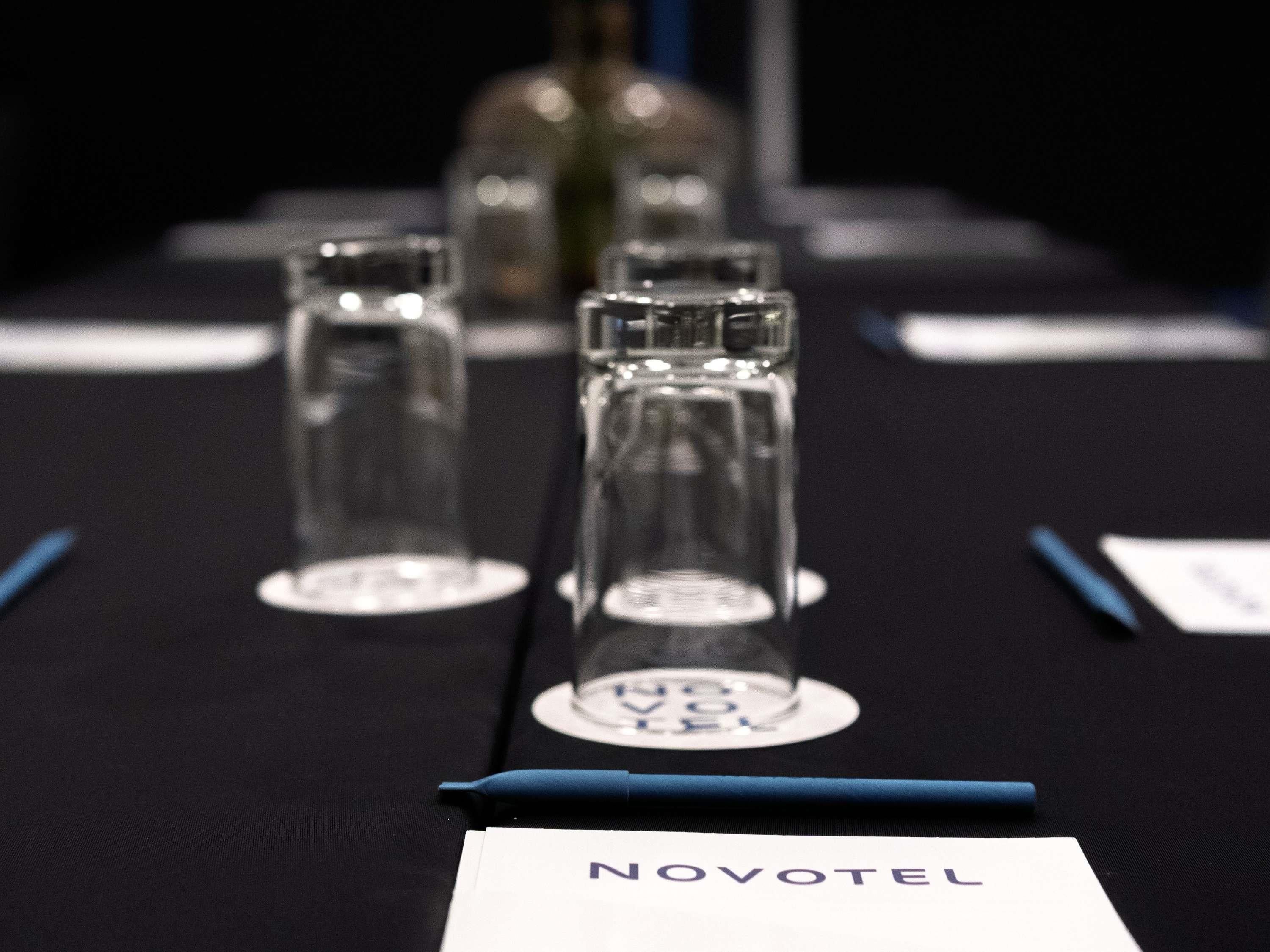 A table with three clear glasses and a pen, a name card with 'NOVOTEL' text, and a blurred bottle in the background.