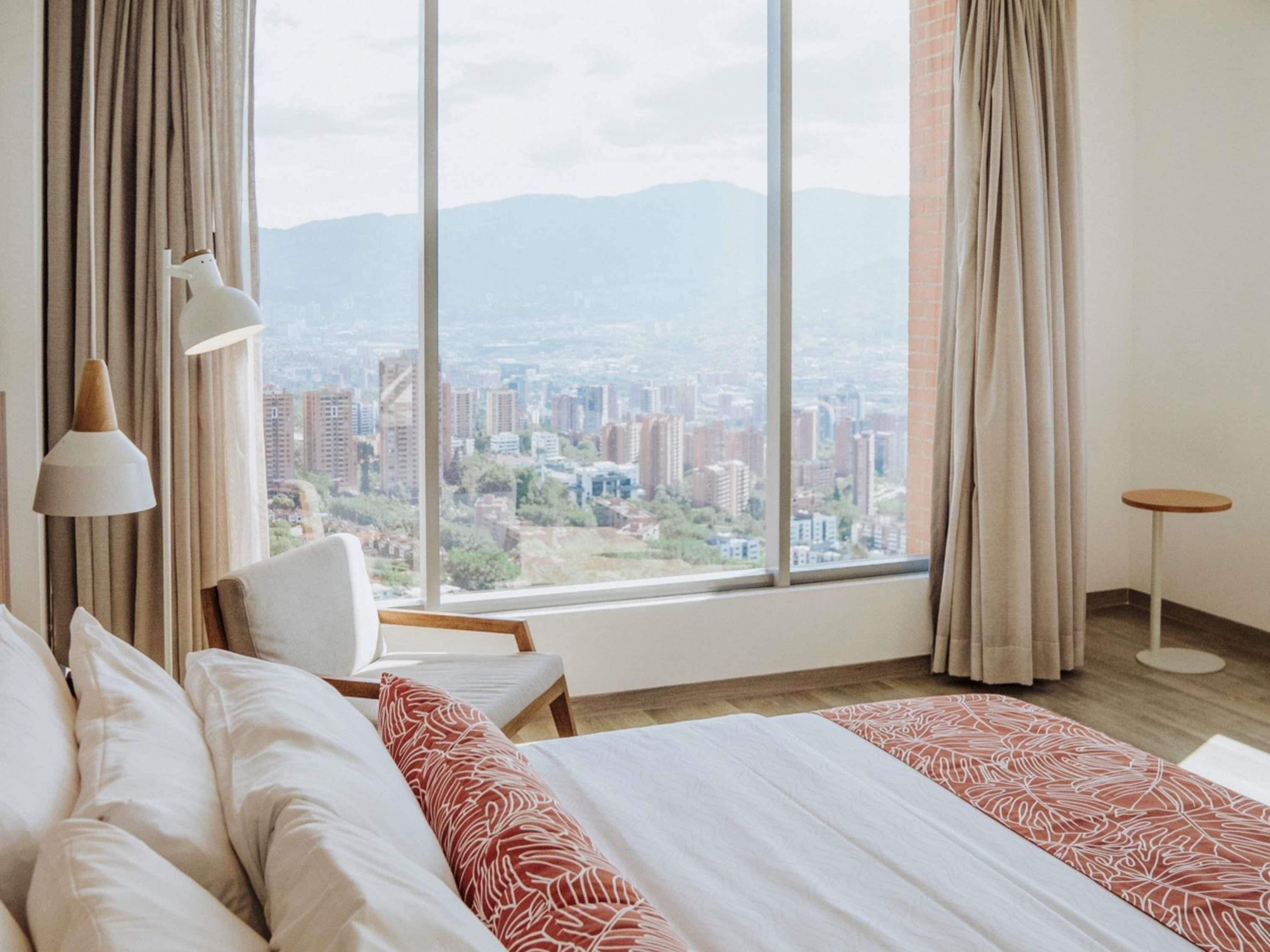 A hotel room with a large window, a bed, a chair, a lamp, and a small table.