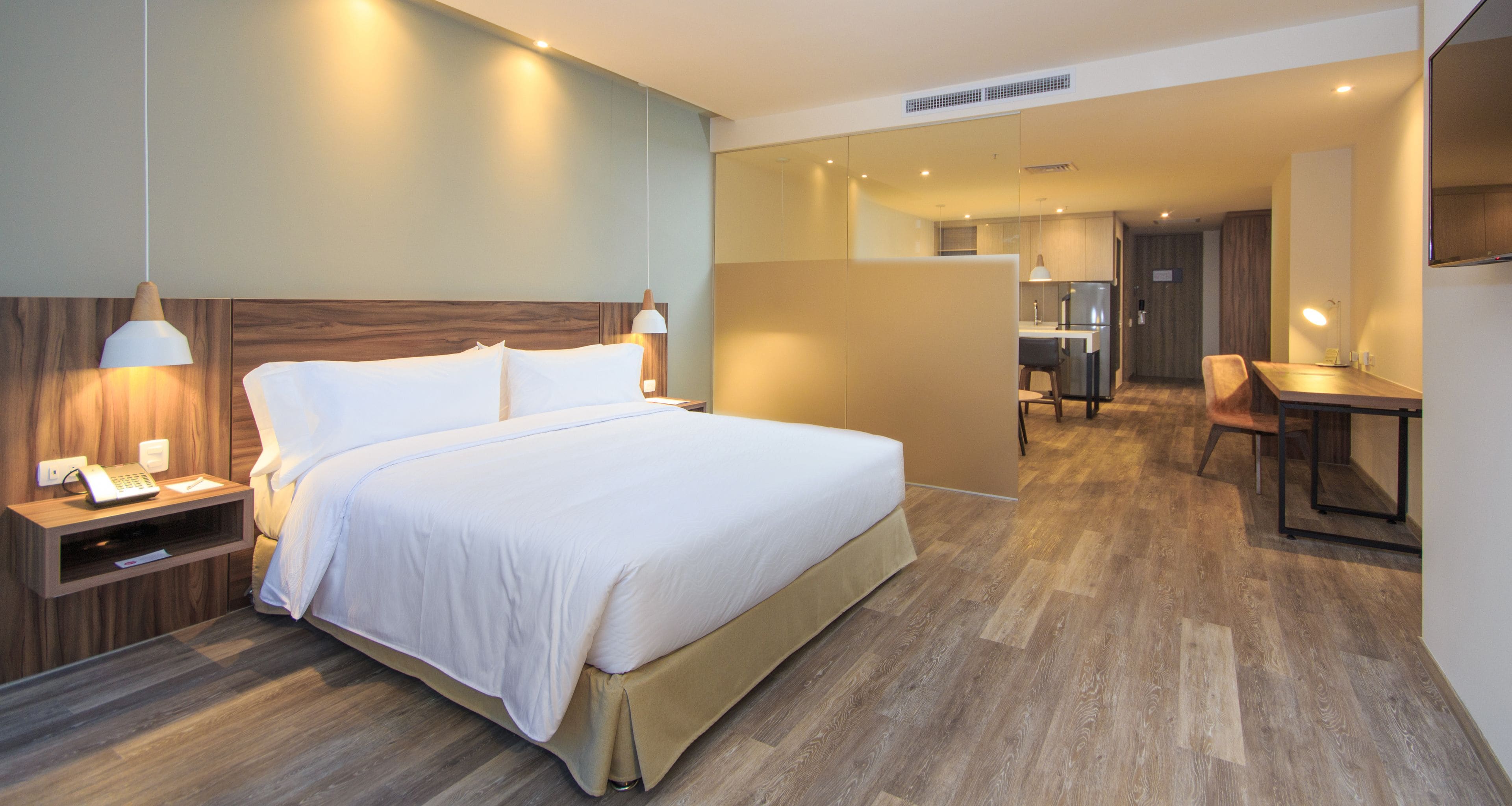 A modern hotel room with a large bed, bedside table with a lamp, a desk with a chair, and a kitchenette in the background.