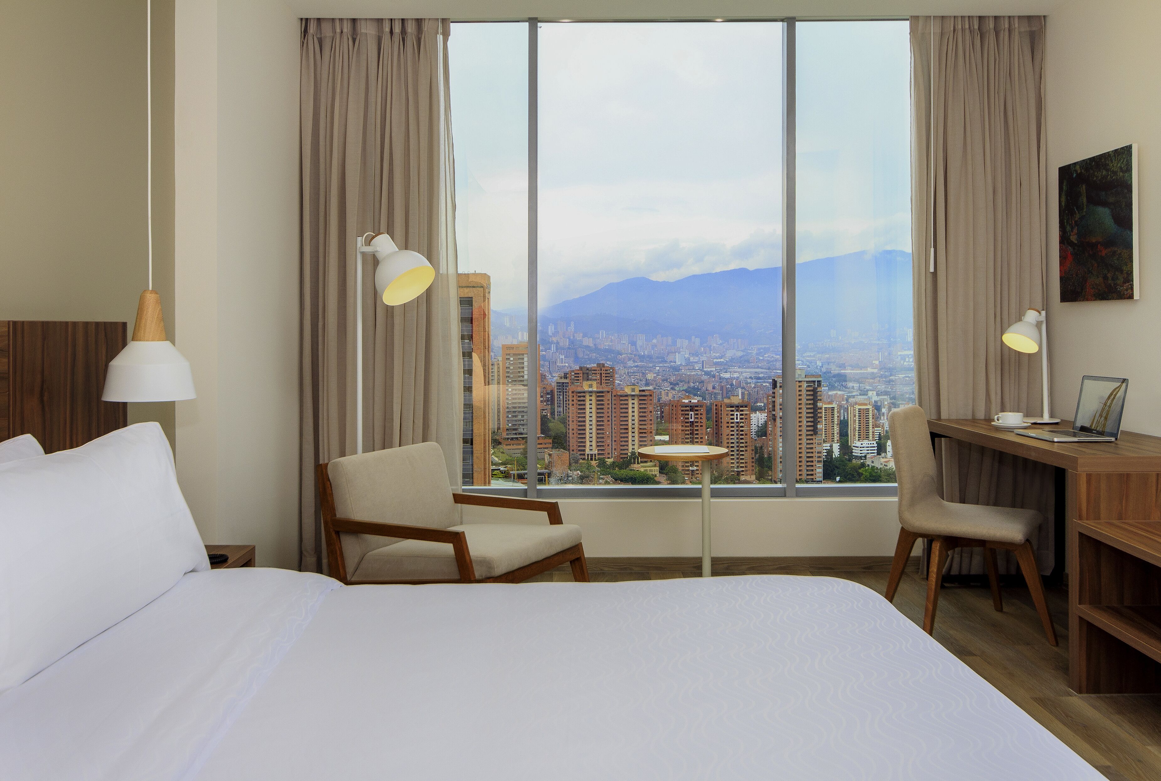 Habitación de hotel con una cama grande, un escritorio, una silla y vistas a un paisaje urbano a través de amplios ventanales.