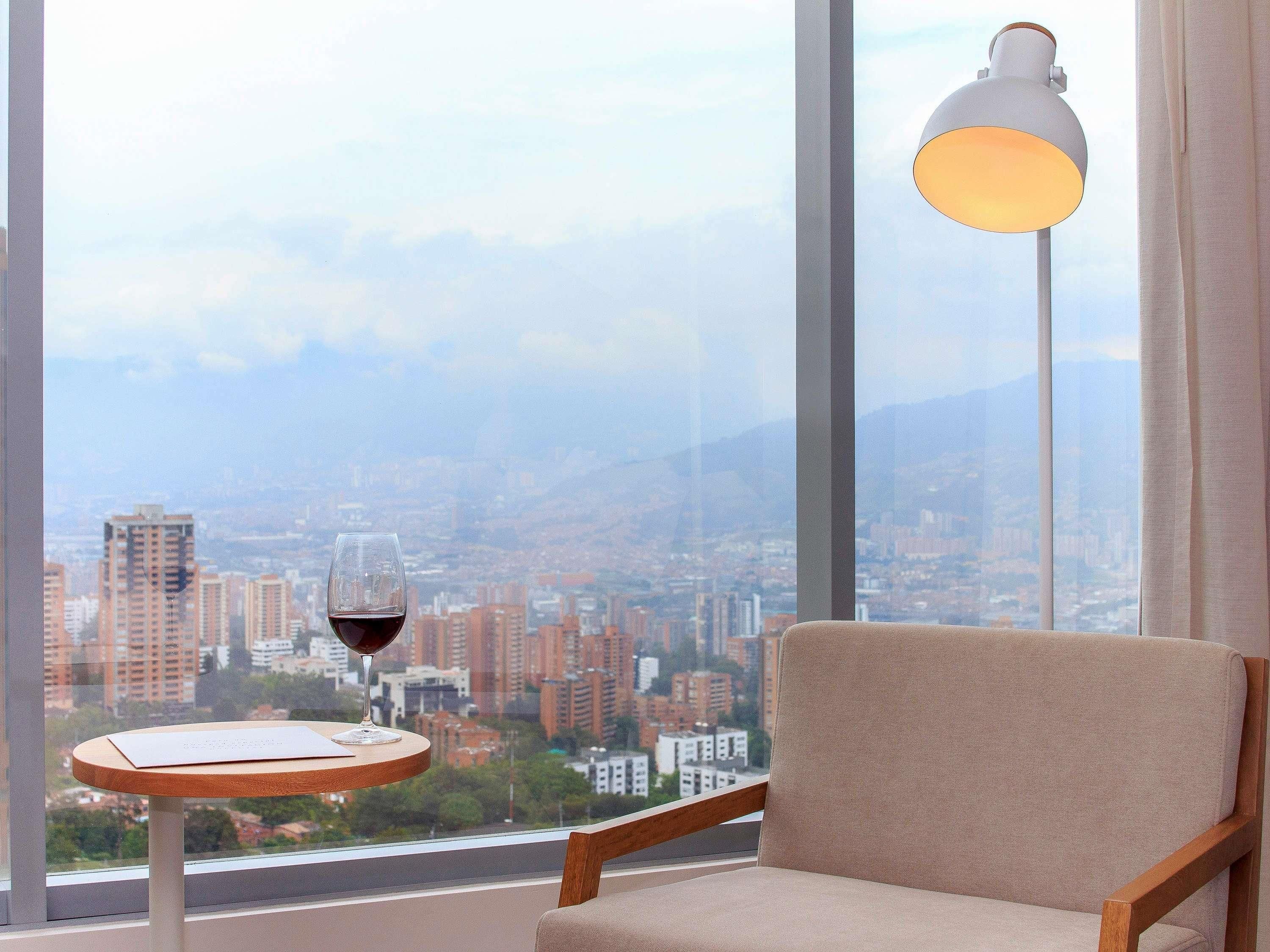 Una habitación con vista a un paisaje urbano, una mesa con una copa de vino tinto y una lámpara.