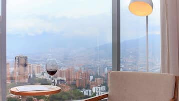 Una habitación con vista a un paisaje urbano, una mesa con una copa de vino tinto y una lámpara.
