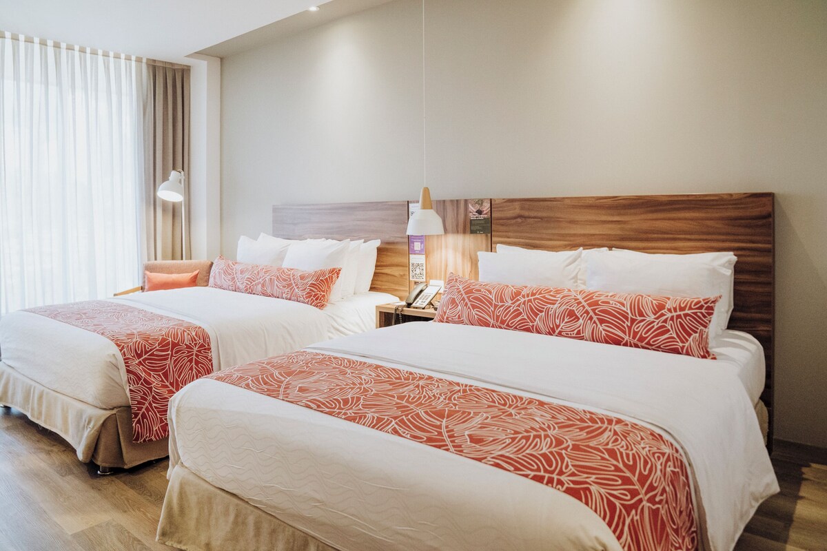 Een hotelkamer met twee bedden, elk voorzien van een houten hoofdbord, wit beddengoed en rode kussens met een patroon. Er staat een nachtkastje met een lamp en een telefoon.