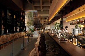 A modern bar with a long counter, shelves with decorative items, and a coffee machine.