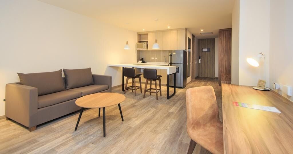 A modern living room with a sofa, a small round table, and two bar stools. There is a kitchen area with a refrigerator, microwave, and a small island with bar stools. The room has wooden flooring and a wooden table with a lamp.