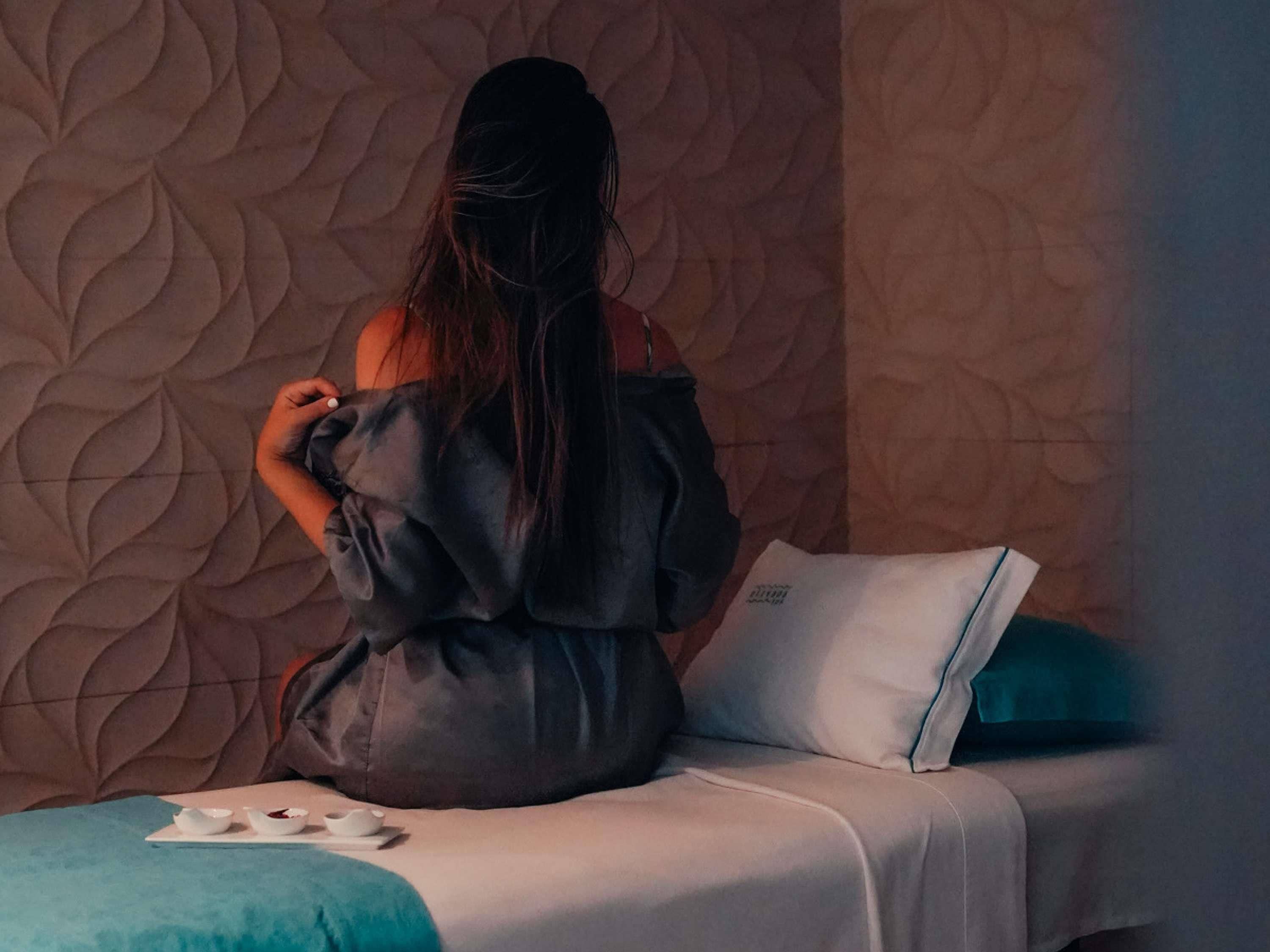 Una persona sentada en una cama con un papel tapiz estampado al fondo.