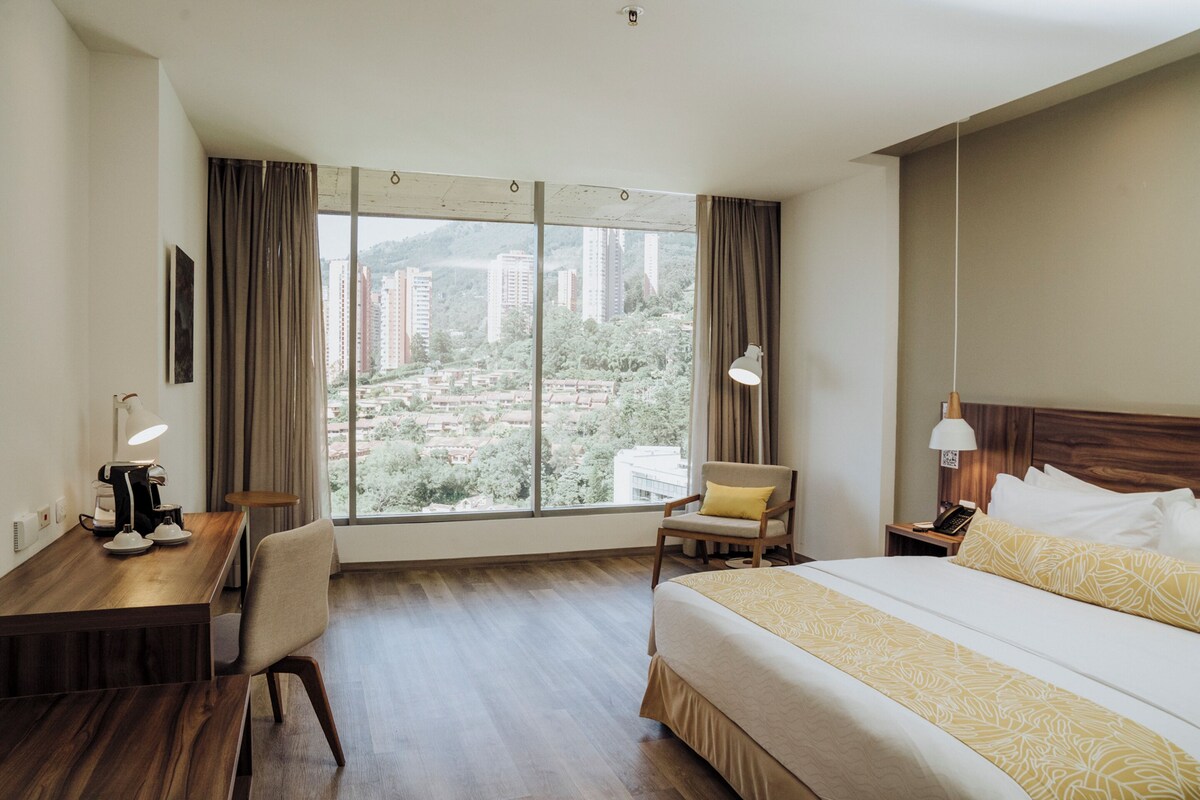 Een hotelkamer met een groot raam, een bed, een bureau met een lamp en een stoel.