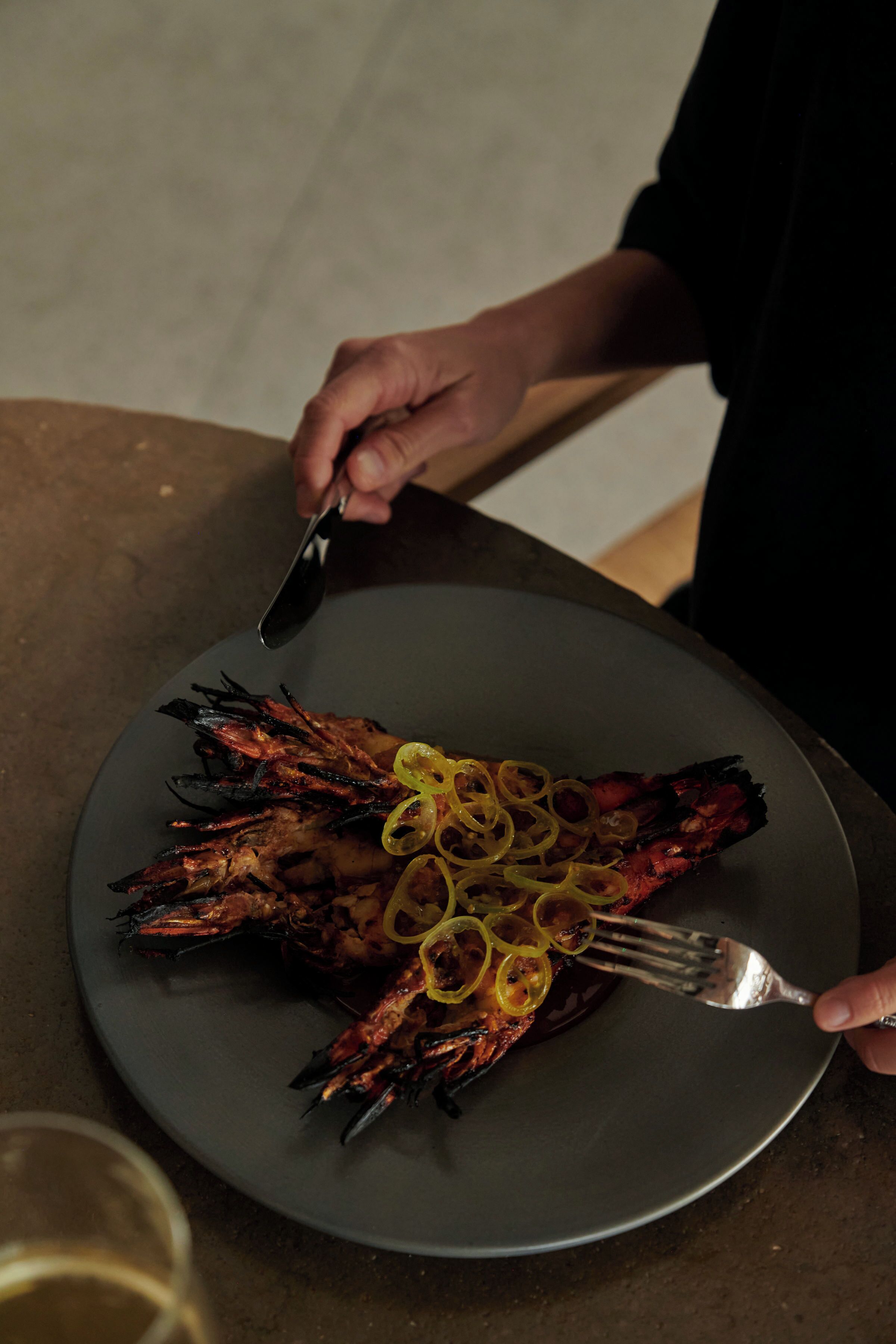 A person holding a fork and knife over a plate with grilled food and lemon slices.