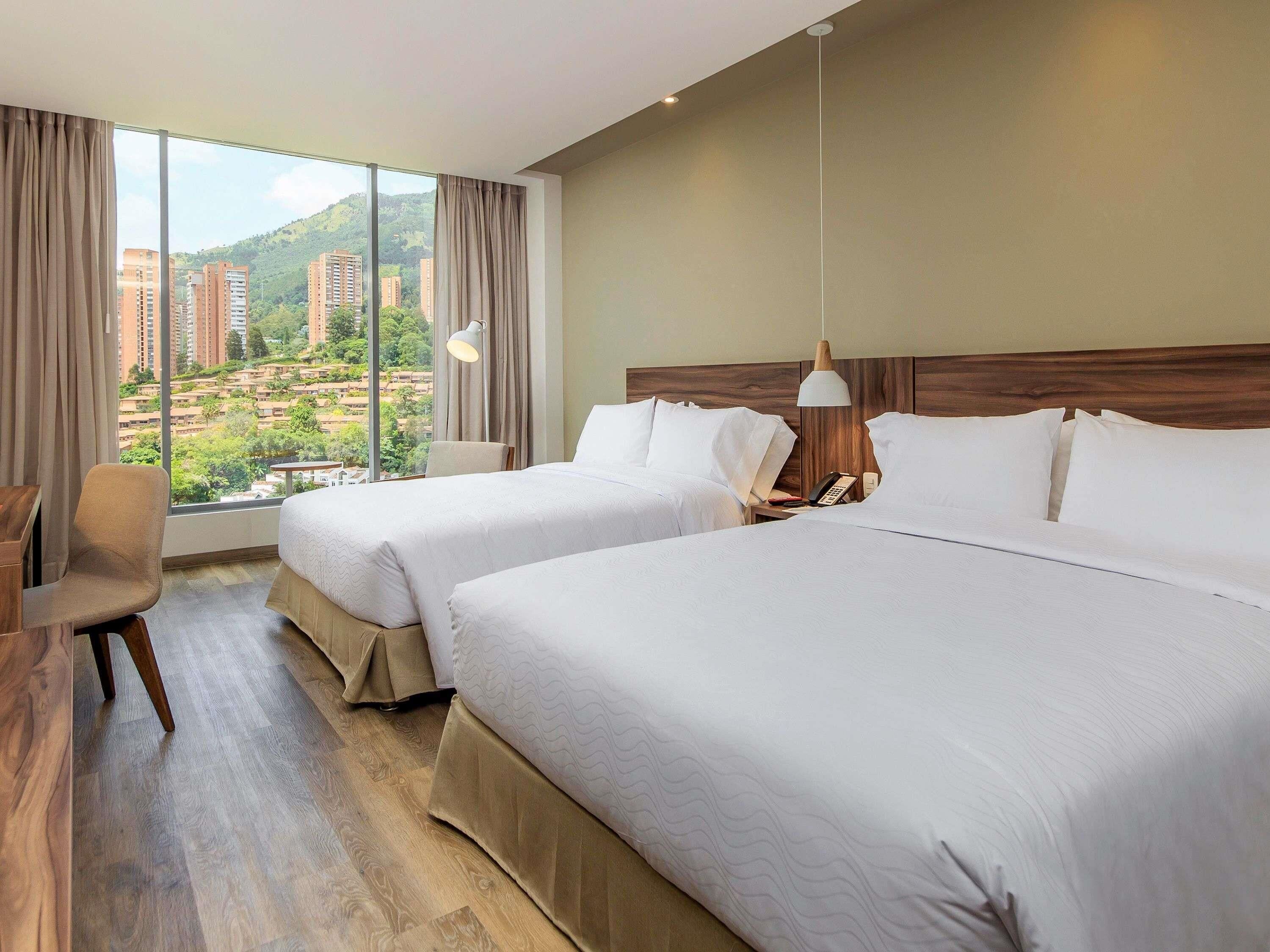Ein Hotelzimmer mit zwei Betten, einem Schreibtisch, einem Sessel und Blick auf Gebäude und Grünflächen.