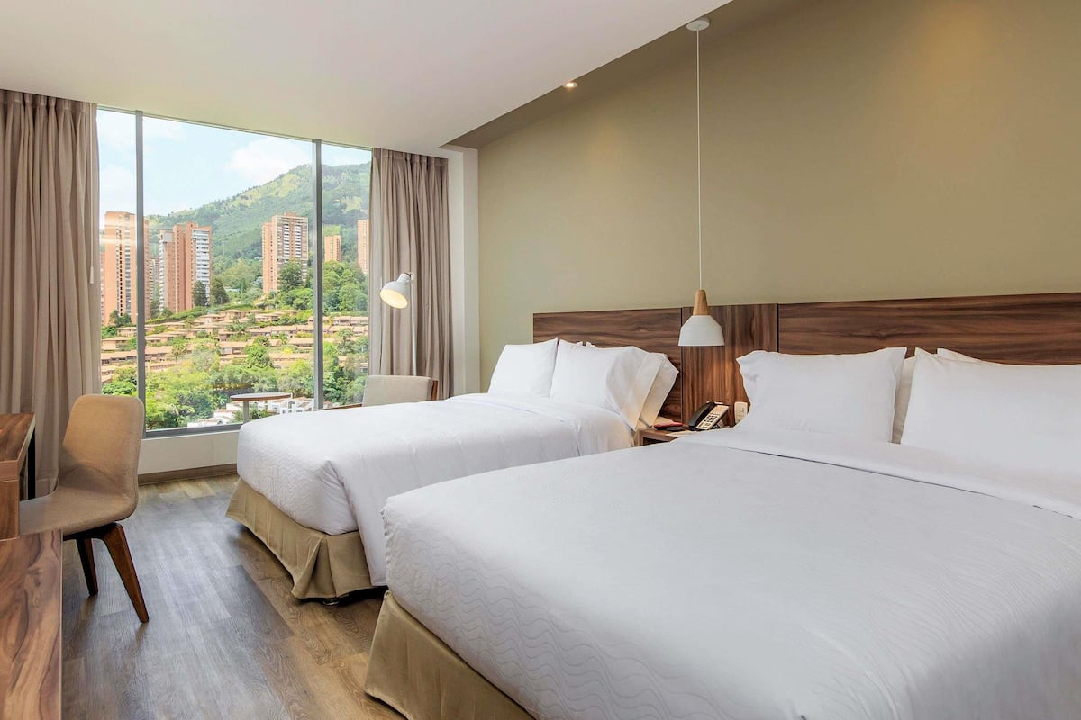 A hotel room with two beds, a desk, a chair, a lamp, and a view of buildings and greenery.