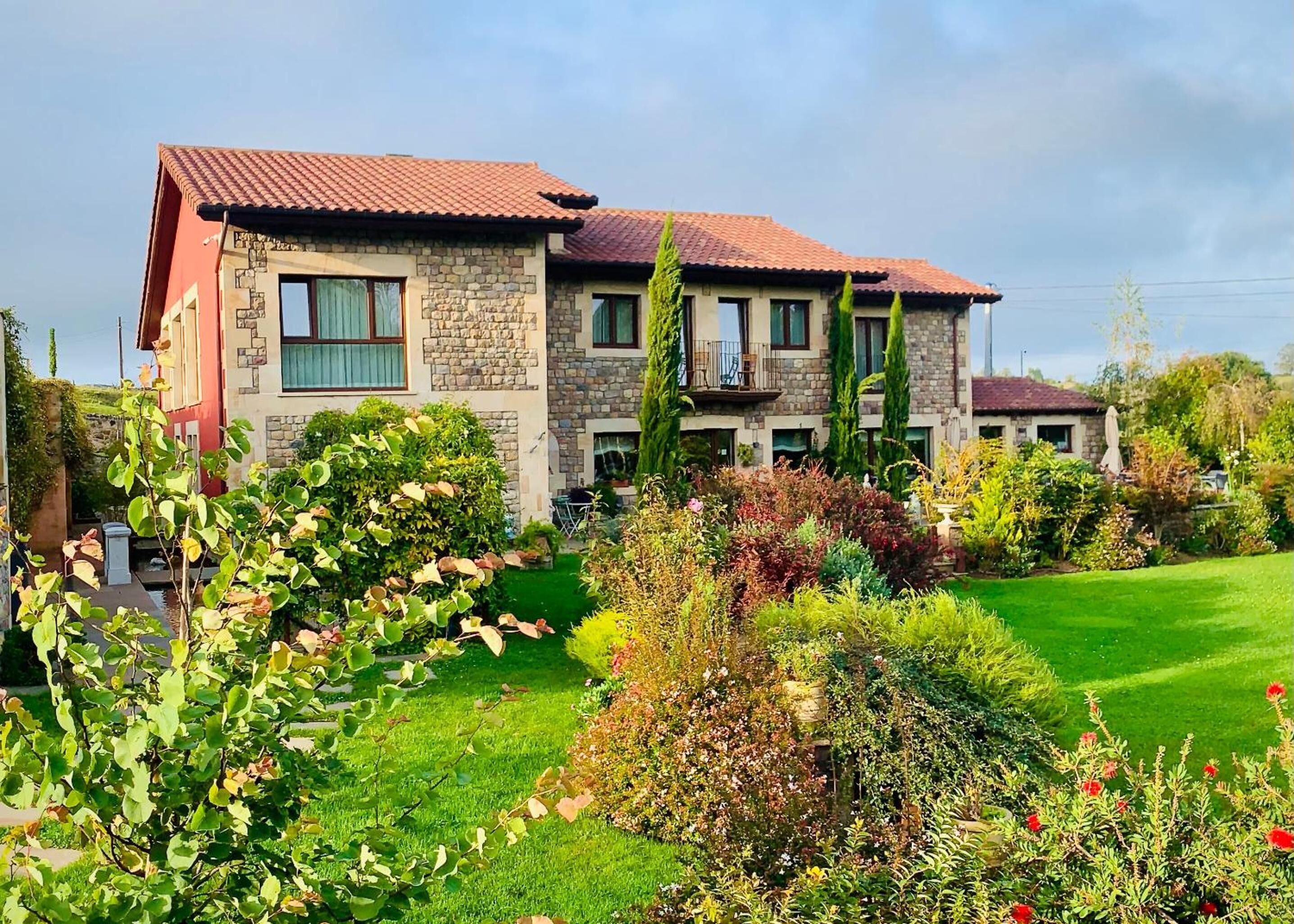 A two-story stone house with a red-tiled roof, surrounded by a well-maintained garden with various plants and flowers.