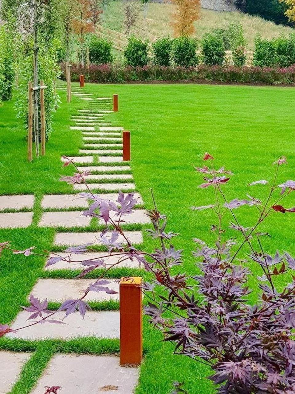 A garden pathway with stepping stones leading through a grassy area, bordered by shrubs and trees.
