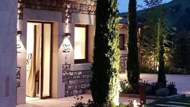 Un edificio de piedra de dos pisos con un balcón, una entrada bien iluminada y un jardín ajardinado con un árbol alto y arbustos.