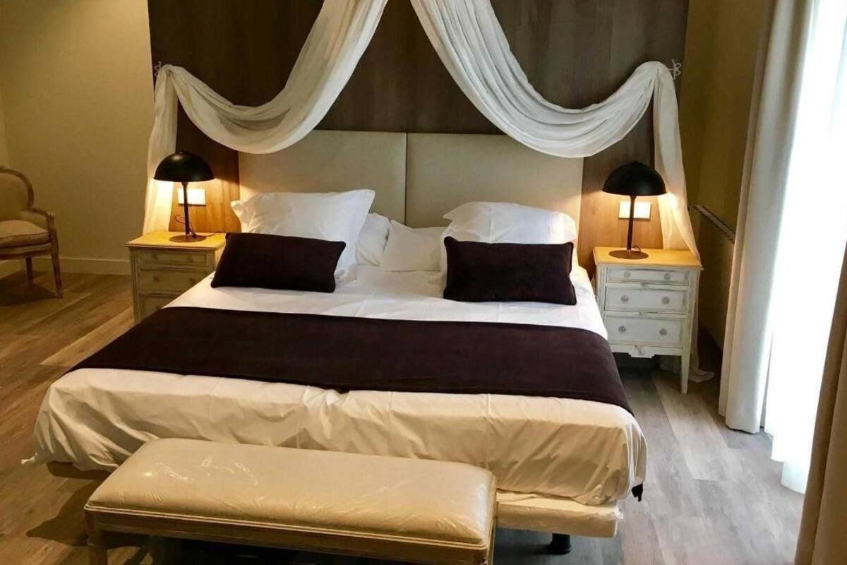 A neatly made bed with a canopy, two bedside tables with lamps, a chair, and a bench.
