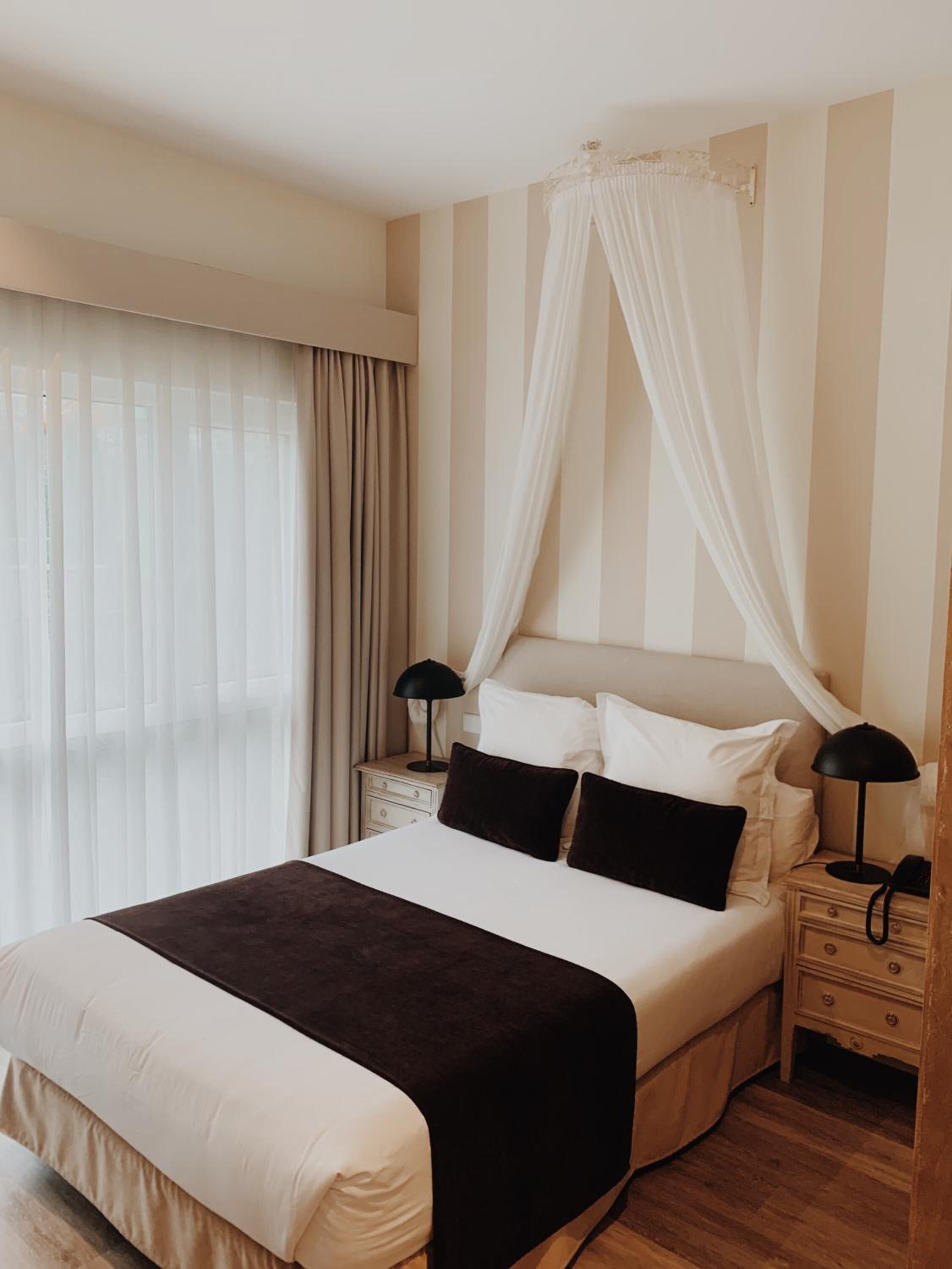 Hotelkamer met een groot bed, hemelbed, twee nachtkastjes met lampen en een groot raam met gordijnen.