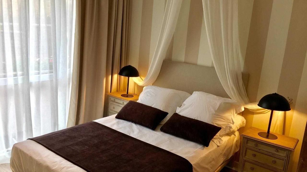 A neatly made bed with white linens and two dark brown pillows, flanked by two bedside tables with lamps, in a room with sheer curtains and a striped wall.