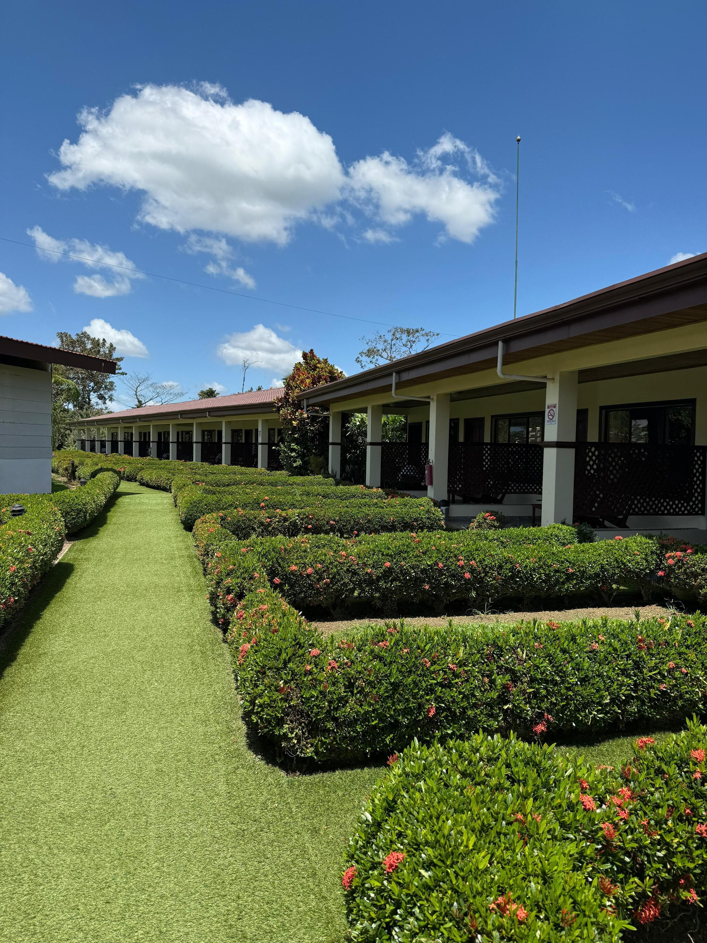 A hotel with a central pathway flanked by neatly trimmed hedges and a row of rooms with balconies.