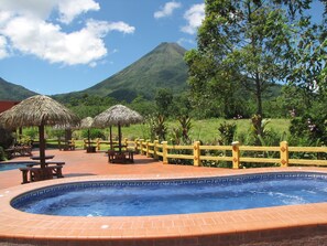 A hot tub area with a wooden fence and thatched umbrellas, surrounded by lush greenery and a mountain in the background. - Hotel La Pradera del Arenal (La Fortuna)