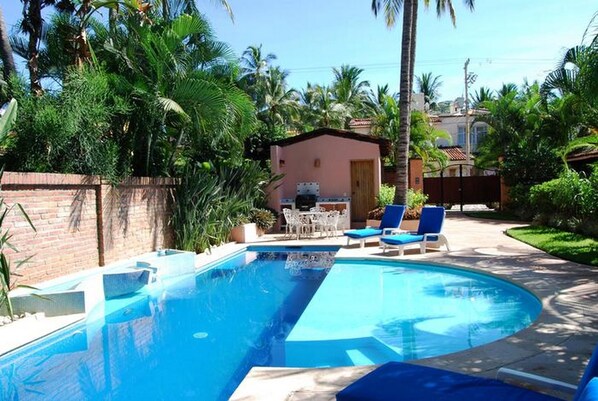 Ein Swimmingpool mit blauem Wasser, umgeben von Palmen und einer Ziegelmauer. Liegestühle und ein kleines Gebäude mit einer Tür sind vorhanden.
