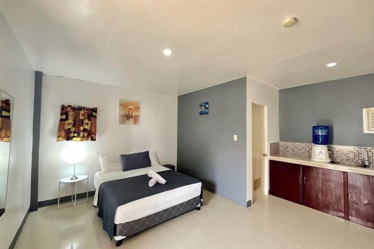 Quarto de hotel com cama, uma pequena mesa de cabeceira, luminária, dispensador de água e uma área de cozinha compacta.