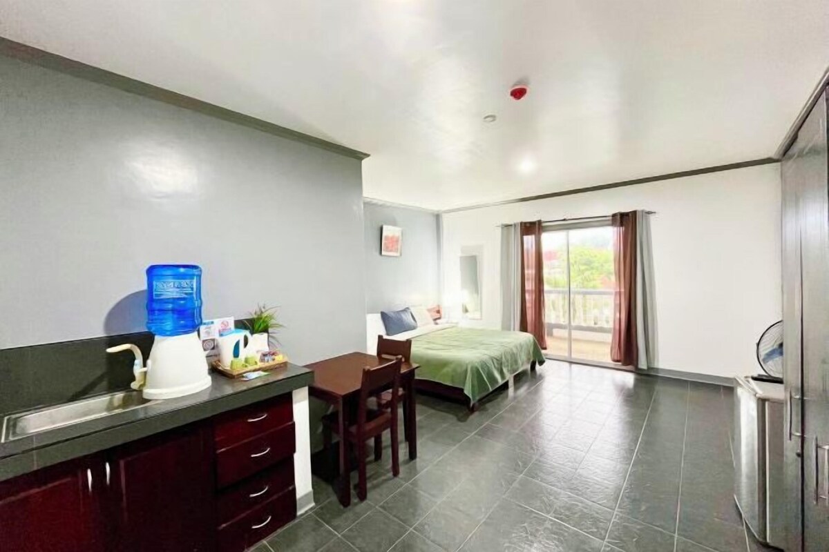 A hotel room with a kitchenette, a bed, a desk, and a large window.