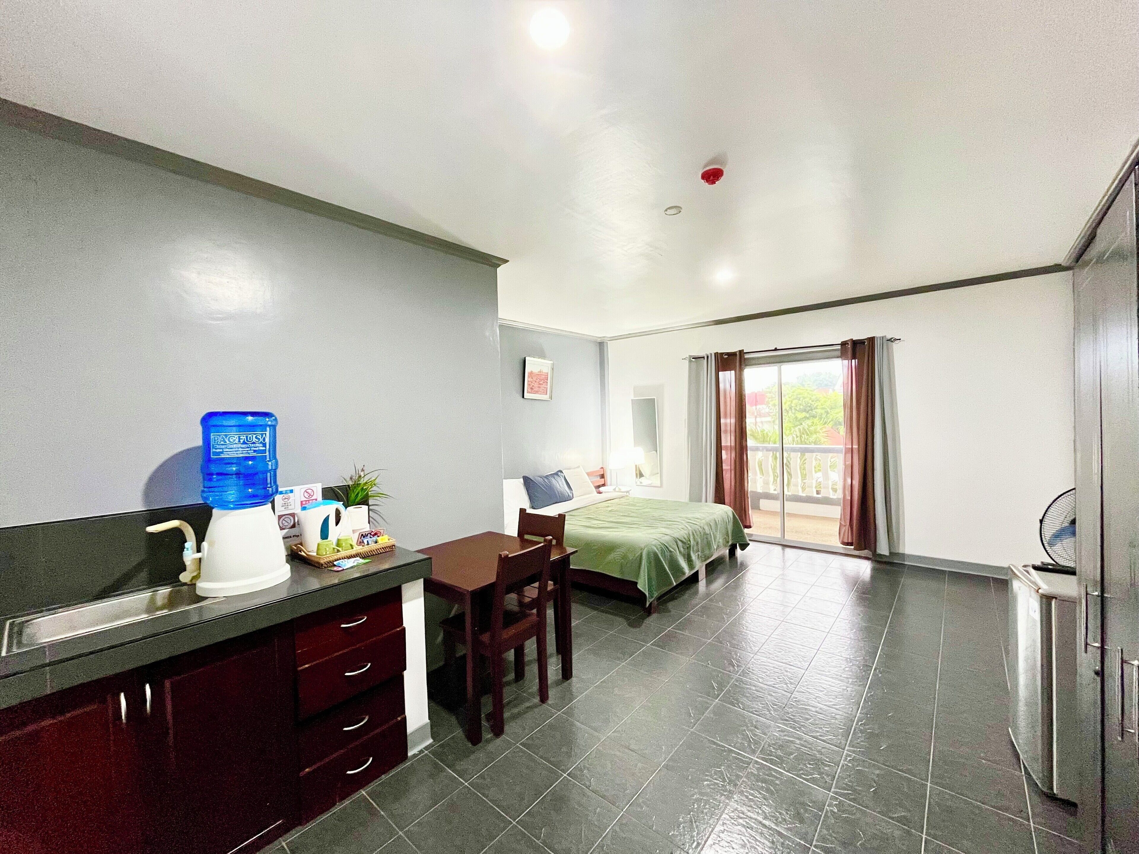 A hotel room with a kitchenette, a bed, a dining area, and a balcony with curtains.