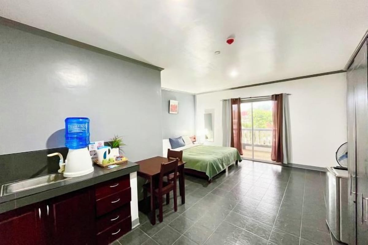 Quarto de hotel compacto com cozinha americana, uma cama, uma mesinha e uma janela com cortinas.