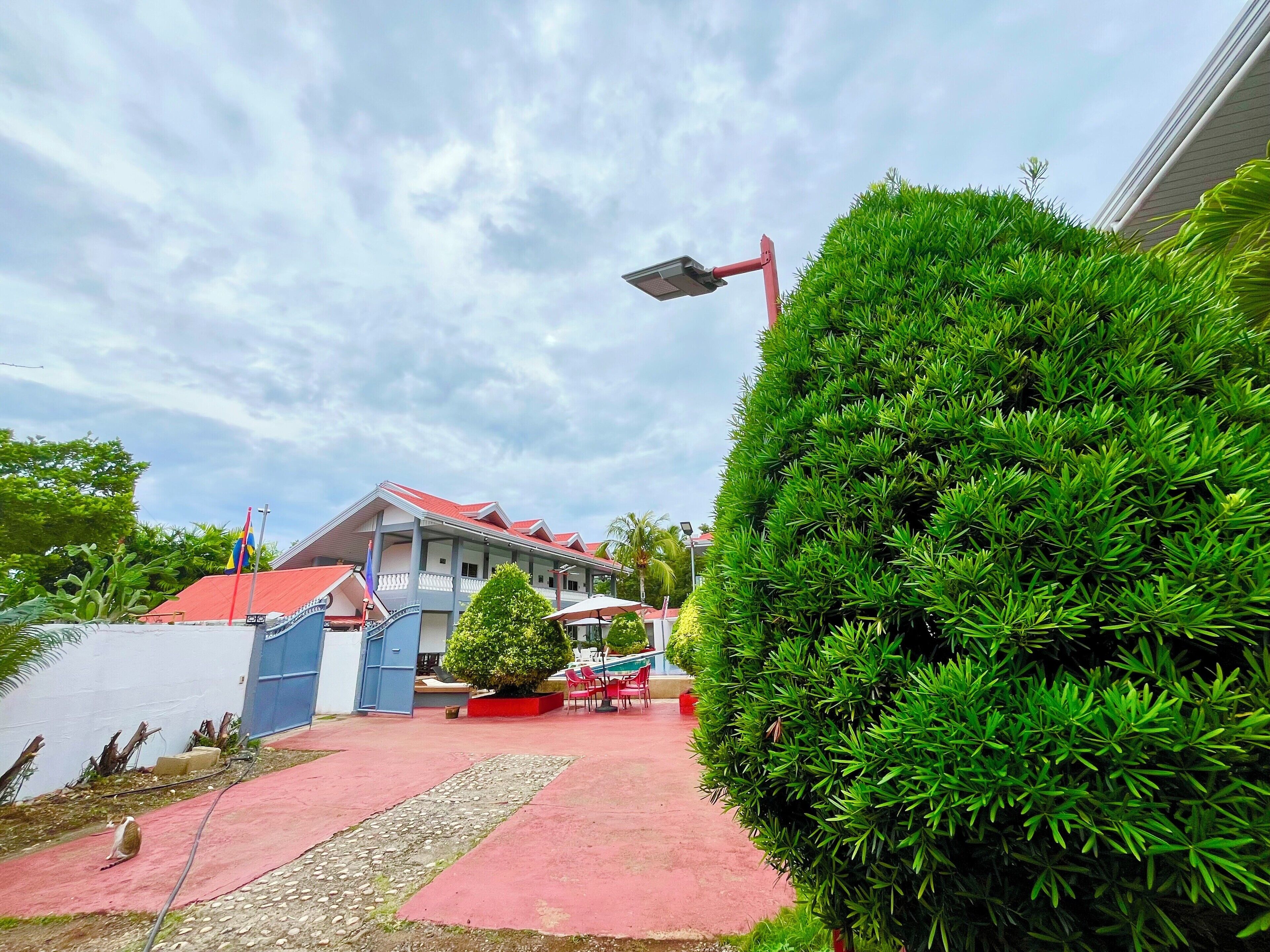 Un edificio con techo rojo y fachada azul, rodeado de setos verdes y un sendero pavimentado.