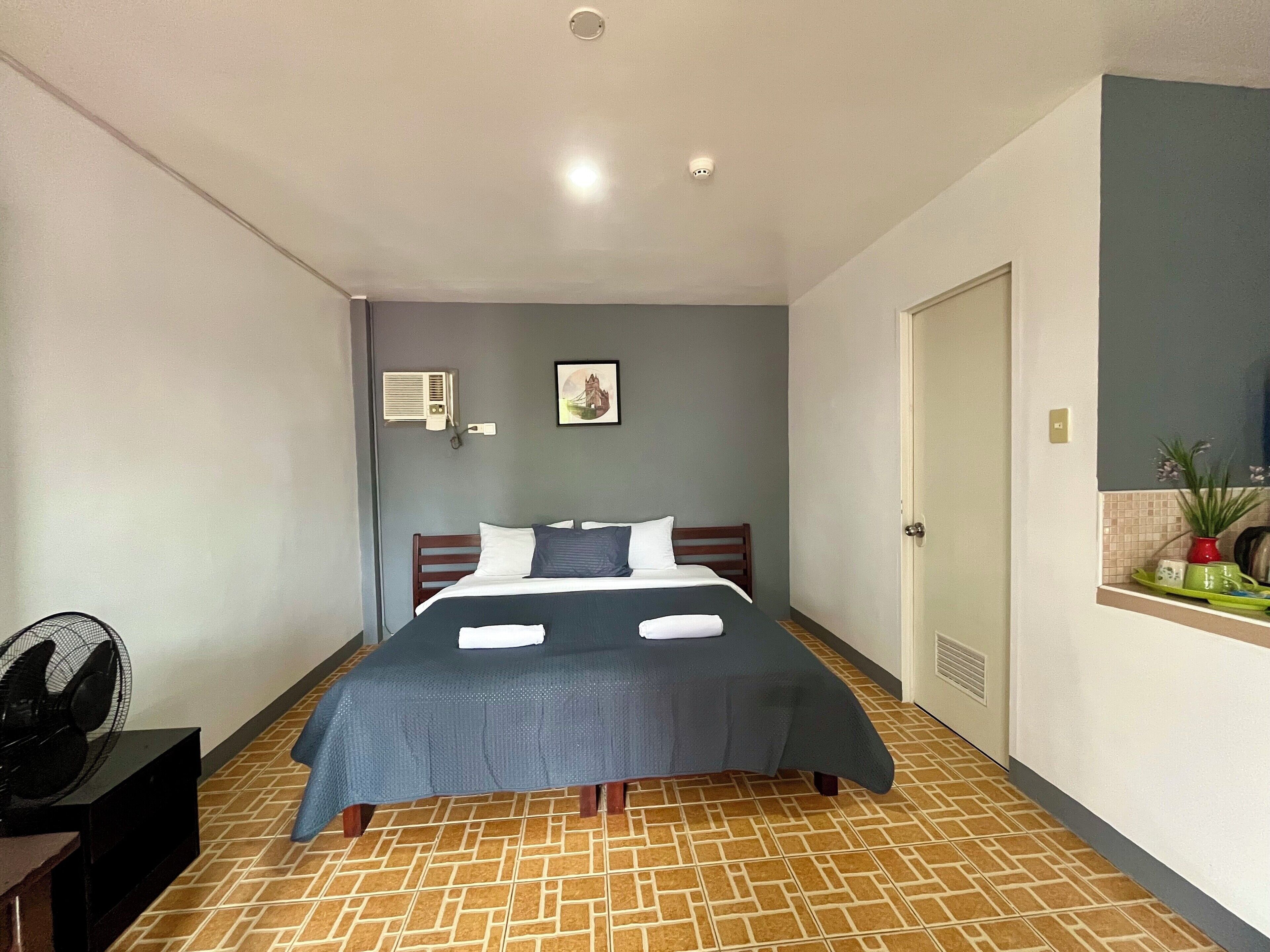 Une chambre bien rangée, avec un lit, un climatiseur fixé au mur, un tableau et une petite étagère ornée d’objets décoratifs.