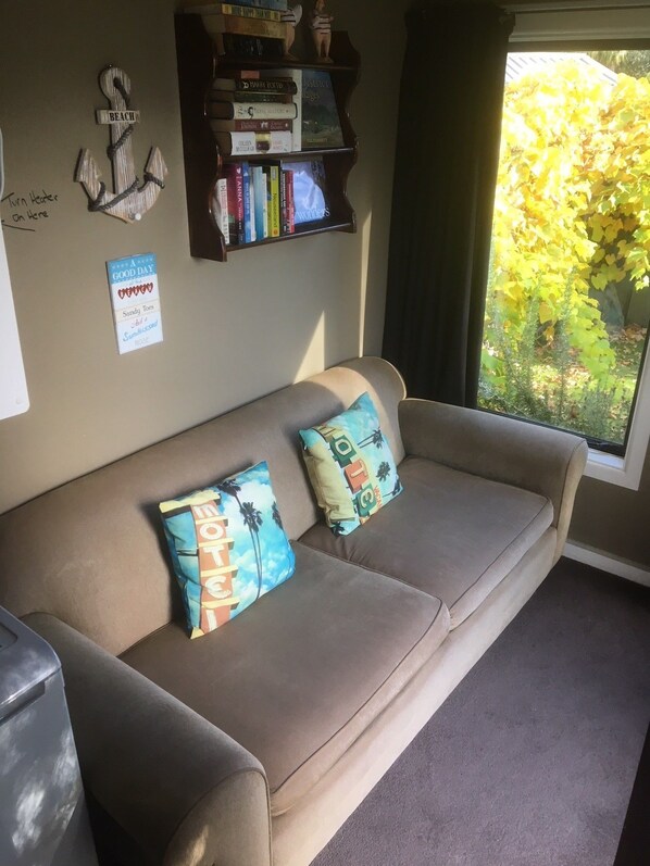 Un sofá color beige con dos almohadones estampados, una estantería con varios libros, un ancla decorativa y una ventana con vistas a árboles.