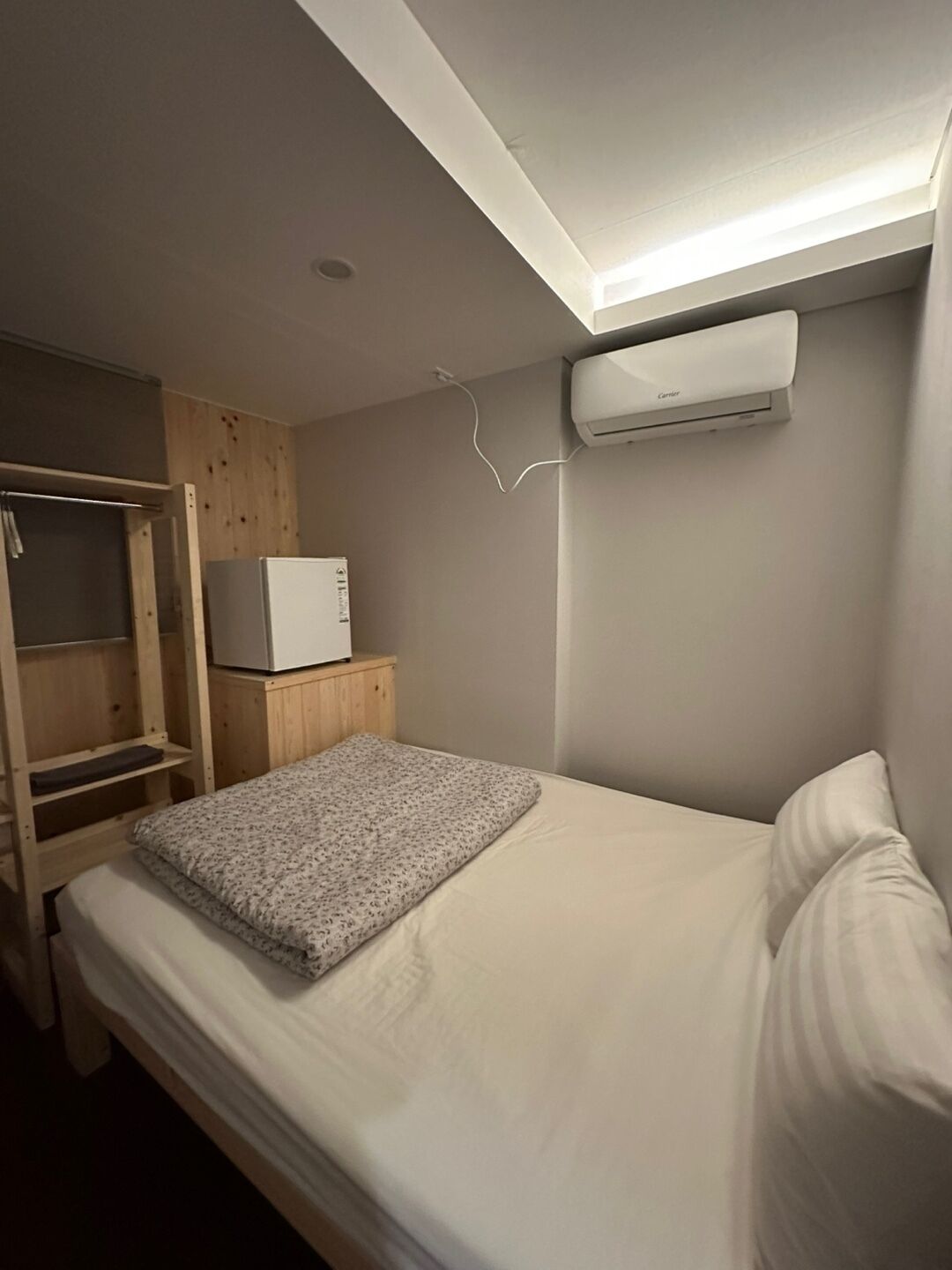 A small, compact room with a bed, a wooden wardrobe, a small table with a microwave, and an air conditioning unit mounted on the wall.