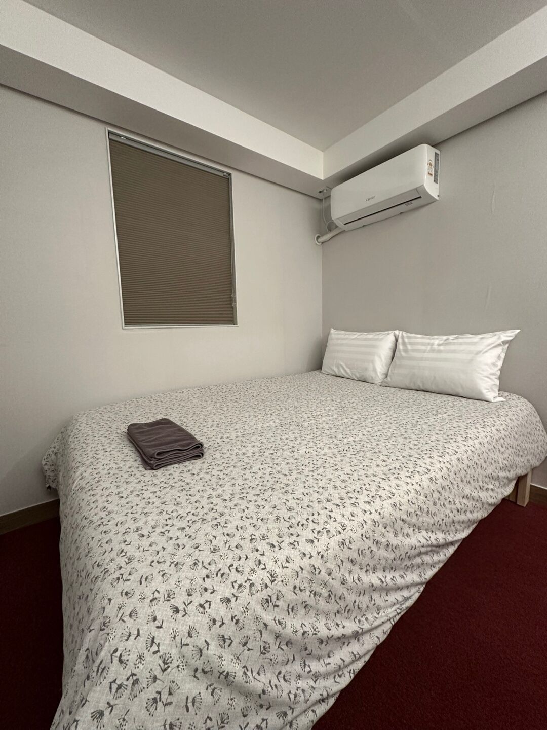 A single bed with a floral-patterned bedspread, a window with closed blinds, and an air conditioning unit mounted on the wall.