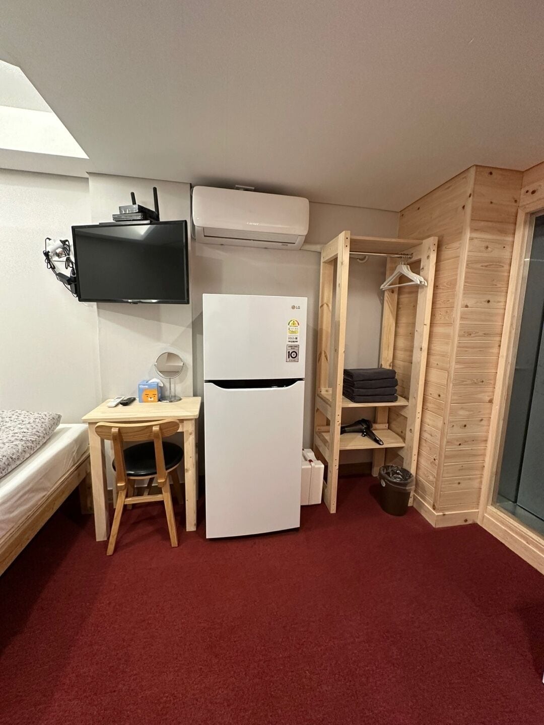 A compact room with a bed, a small desk, a white refrigerator, a mounted TV, and a wooden wardrobe.