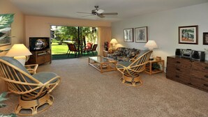 A room with wicker furniture, a television, and a view of a garden through a sliding door. - Hale Kamaole 133 Ground floor condo, easy access to best beach on Maui! (Kihei)